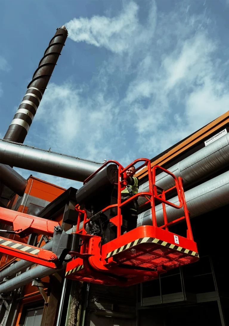 Servicetekniker i skylift utför arbete på industribyggnad med ventilationsrör, framför en fabriksskorsten under blå himmel.