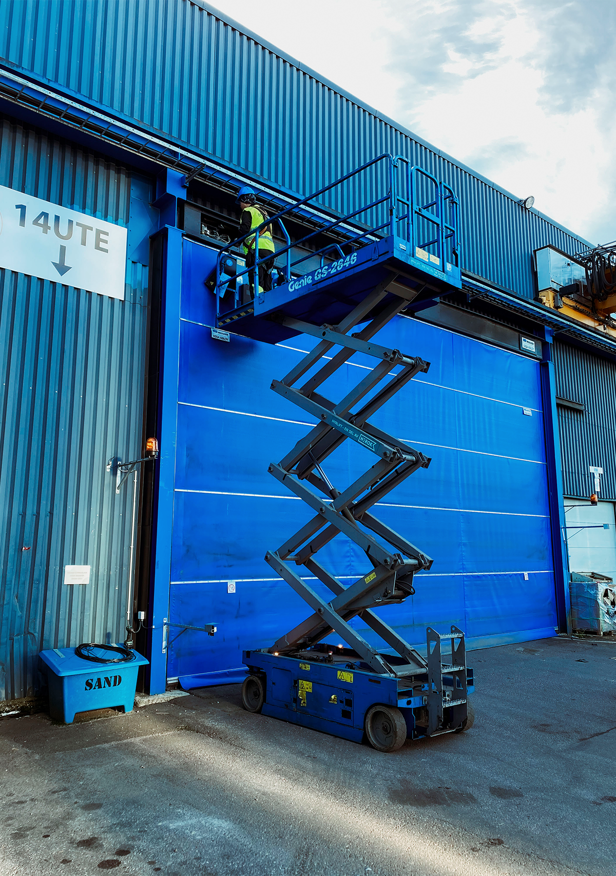 Servicetekniker arbetar från saxlift vid en stor industriport i blått.